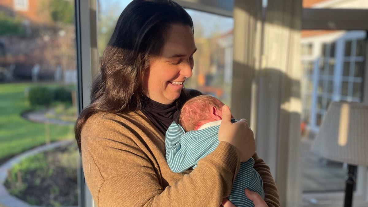 Amy, 36, has long brown hair and is smiling down at the baby she is holding against her chest. She stands in front of a window through which a neat garden can be seen. Sunlight falls on her face and hand. She is wearing a biscuit-colour cardigan and is holding a baby that is a few days old. The baby is wearing a blue and white striped baby-grow. He faces away from the camera and has a wrinkly pink head and sparse hair.