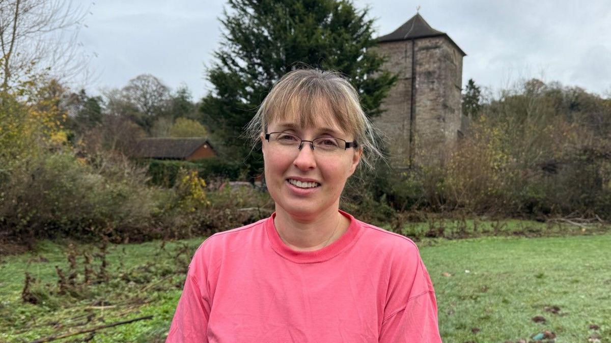 A woman in a pink top looking at the camera with a building in the background, which is mainly grass, trees and sky.