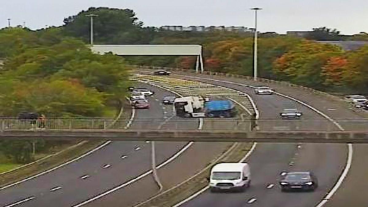A lorry cab in the middle verge between two double lanes of traffic with a footbridge in the foregound, autumn trees behind and several vehicles driving by