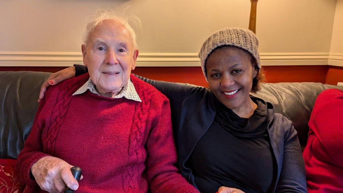 A woman wearing a black top and jacket sitting on a brown leather sofa with an elderly man wearing a red knitted jumper. She has an arm around his shoulder and they are holding hands.