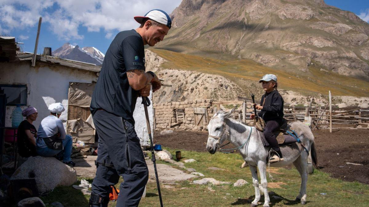 Mr Jackson standing outside what appears to be a local shepherd's house at the base of a mountain. It is a small white shack with a wooden door and roof, and a wooden fence around the perimeter. He is wearing black trousers and a black T-shirt, with a blue and white cap on backwards. He is standing up and leaning on a hiking pole, looking down towards the ground. A few feet in front of him there is a local woman wearing black, sitting on a white horse.