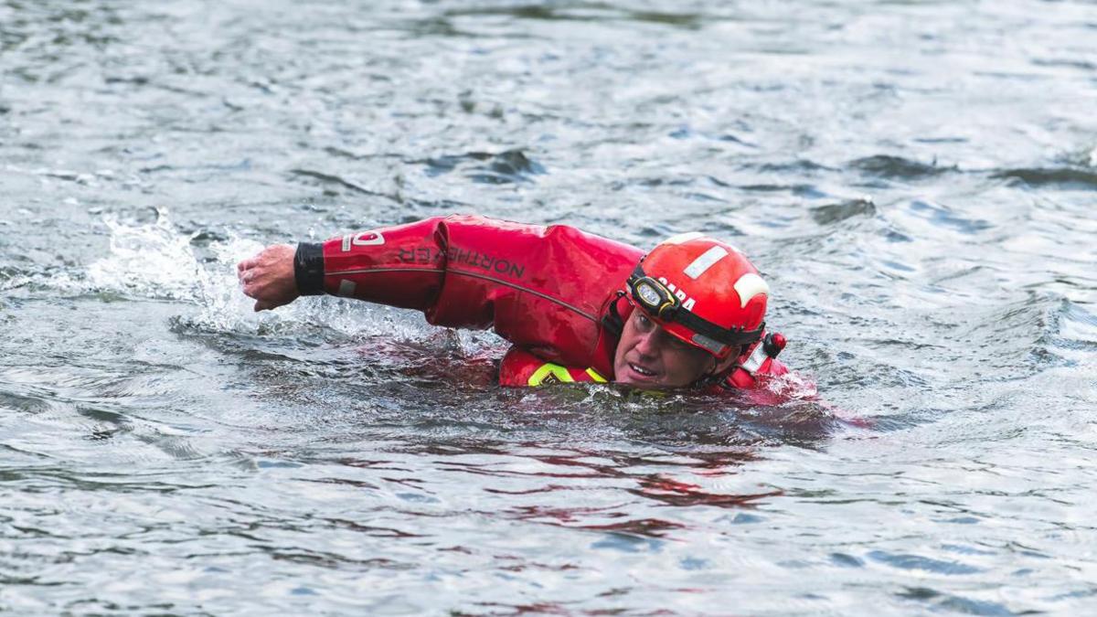 A man is swimming frontcrawl in a river while wearing red overalls and a red helmet, which has front light attached to it.