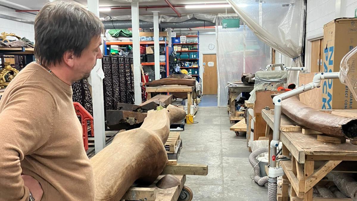 A man wearing a brown shirt from the conservator team, standing in a studio with a long piece of the arch in front of him. He is inside a workshop with various benches and tables and equipment. Plastic sheet curtains are pulled back o nthe right side. There is a door in the distance with a clock above it.