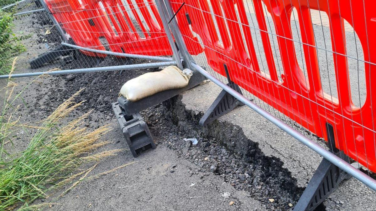 A close up of broken tarmac on the pathway. It is blocked off by red safety fencing and silver fencing. 