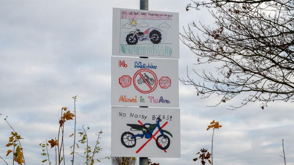 Three white signs on a metal pole. The top sign depicts a bike with big black tyres driving on grass. The sign below it has a man riding a bike with a red line through it and two red 'stop' signs. The bottom sign has a bike with a red cross drawn over it