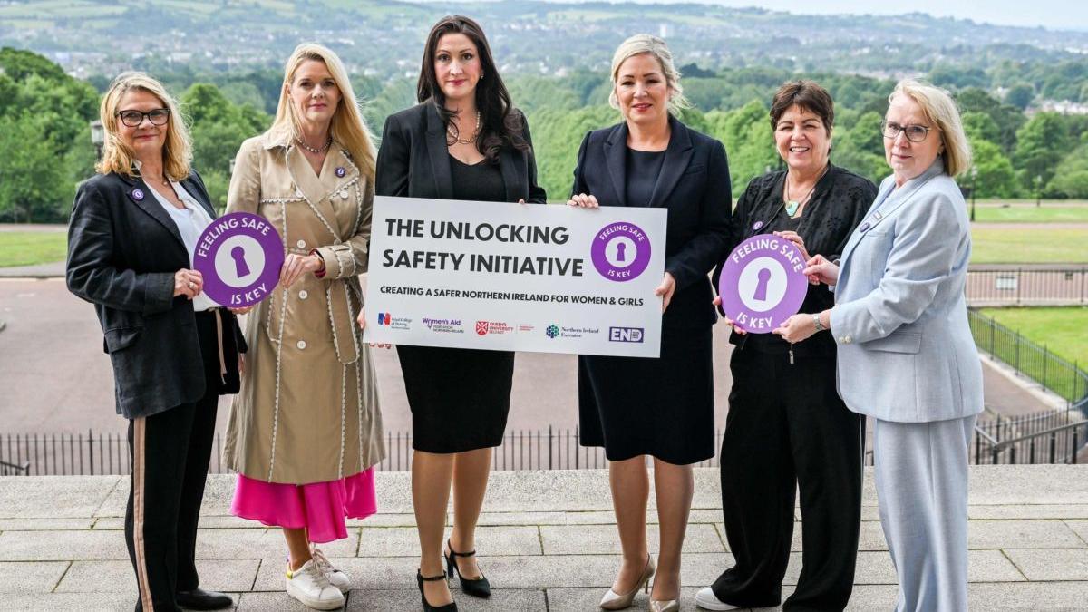 Representatives from Women's Aid, QUB, the RCN and the Executive Office stand beside Emma Little-Pengelly and Michelle O'Neill, who are in the middle, holding a sign reading "The Unlocking Safety Initiative - Creating a Safer Northern Ireland for Women and Girls". They are stood outside on the steps outside Stormont.