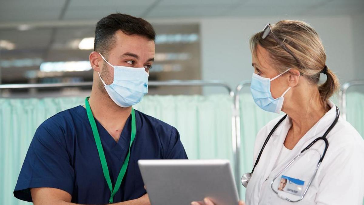 A man with short dark hair and a blue medical top with a green lanyard looks at a woman with tied up blonde hair and a white medical top with a stethoscope around her neck. The woman is holding a tablet and both are stood in a medical ward with pale green curtains behind them. Both are wearing face masks. 
