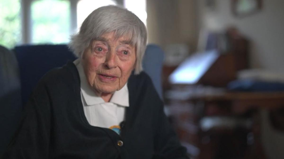 An elderly woman with grey hair sat in an armchair. She is wearing a navy blue cardigan and a white collared shirt.