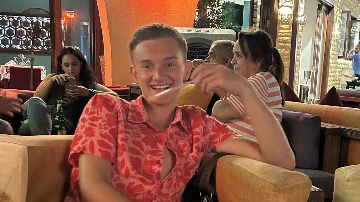 Jack Dolan sits in an armchair in a pink and red shirt. He has short blond hair and is smiling. 