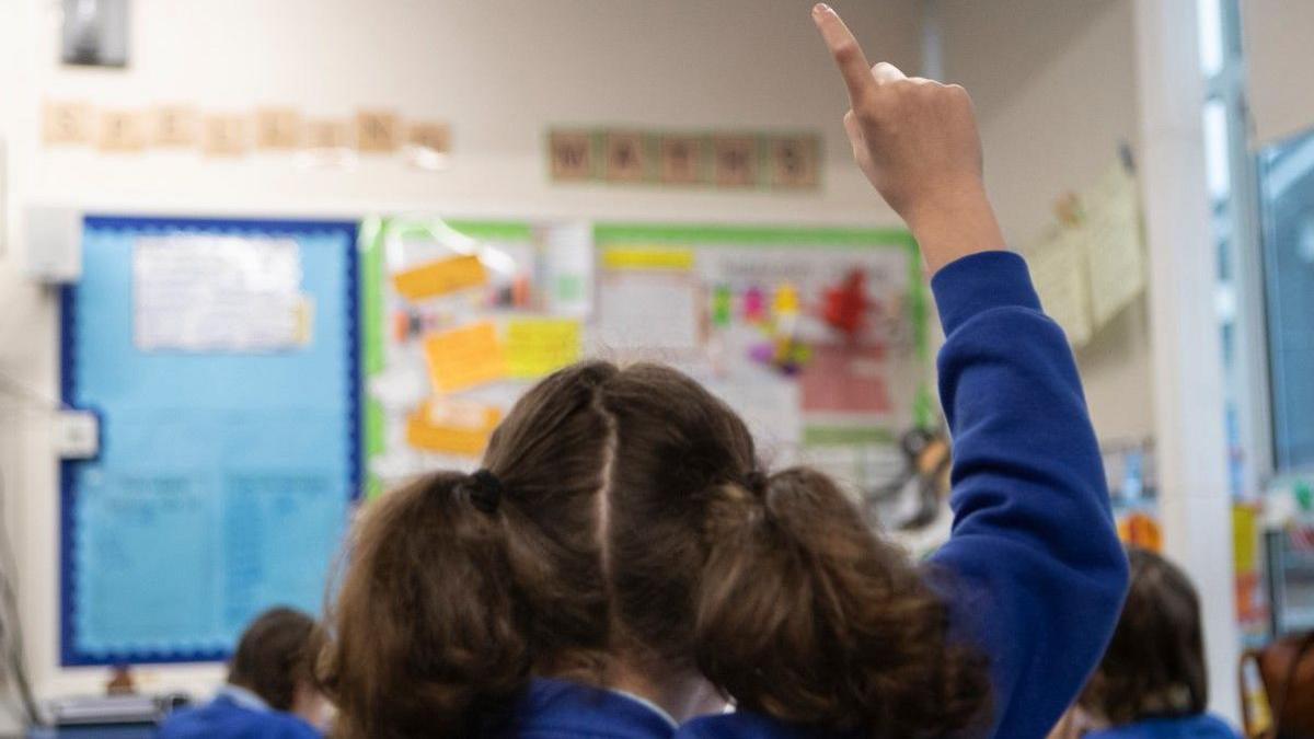 Rear shot of Year Five pupil with hair in bunches and wearing a blue sweatshirt raising her right hand in class.