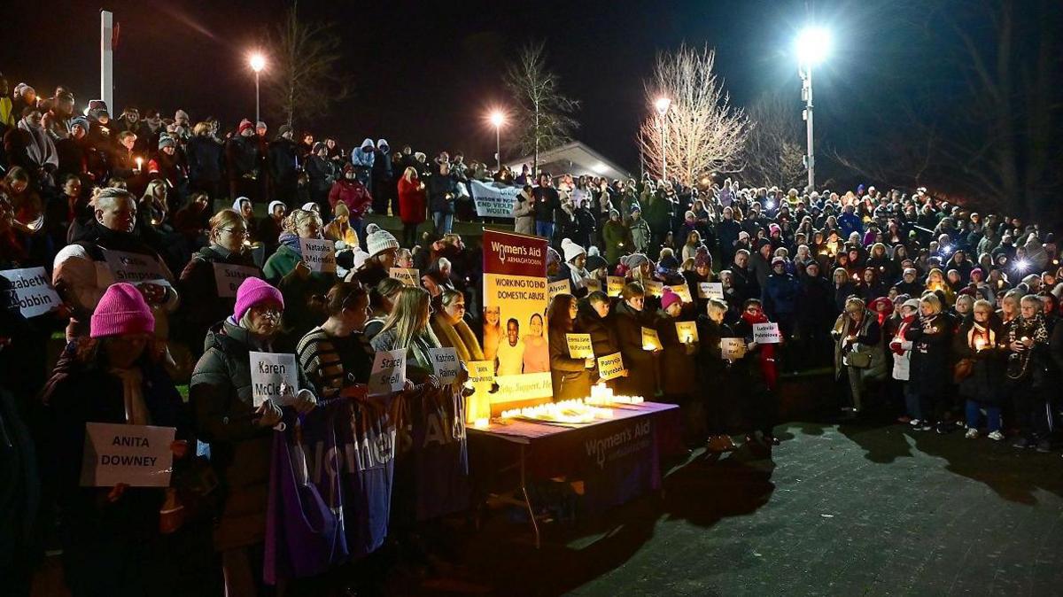 The crowd of mainly women, shown from a side angle. There is a table to the front and in the middle with candles on it and a sign protesting against violence against women.