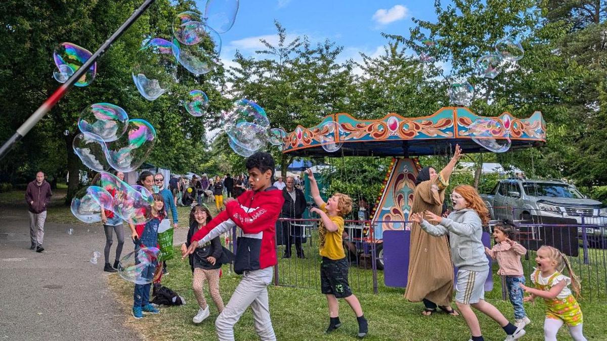 Children are chasing and popping balloons at the festival. In the background is a merry-go-round.