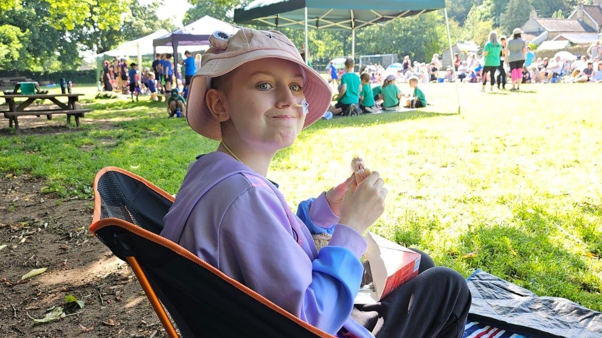 Megan wearing a hat sitting in a chair outside with other children in the background