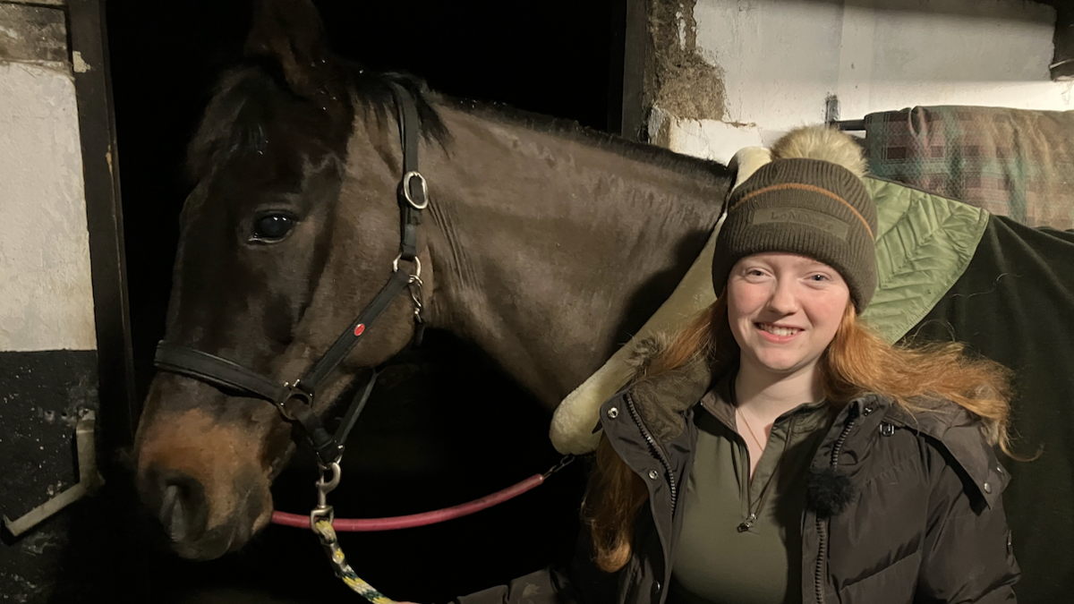 Samantha and horse Ozzy standing in a stable, Samantha smiles at the camera. Ozzy stands behind her, a black horse with a green saddle. 