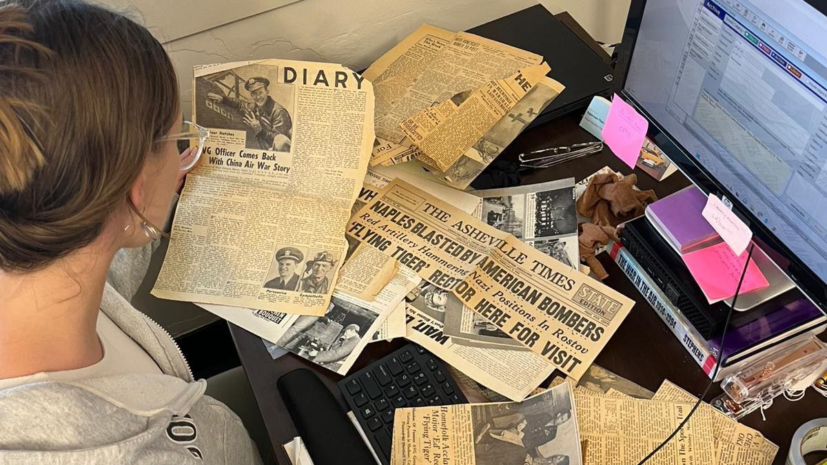 A blonde woman sitting at a computer screen with old newspaper clippings spread out over the desk in front of her.