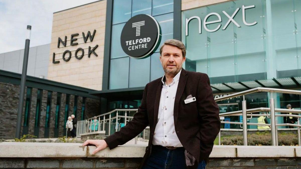 Glynn Morrow is standing in front of the Telford Centre, which is a large glass-fronted building. It has a Telford Centre sign on the front, along with signs for New Look and Next. Mr Morrow has brown hair and facial hair, and is wearing a white shirt and dark burgundy jacket