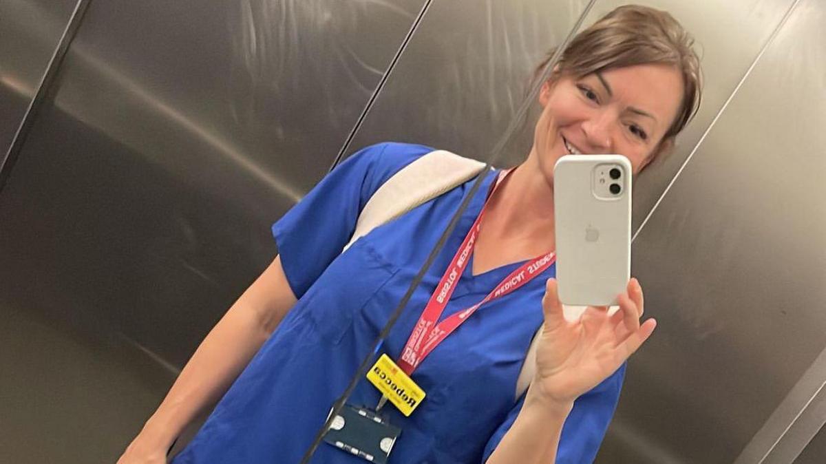 Dr Becs Bradford. She is inside a lift, taking the photo with her phone in the mirror. Her long dark hair is in a ponytail and she is wearing blue scrubs and a red lanyard. She is smiling, although the phone covers up a section of her face. 