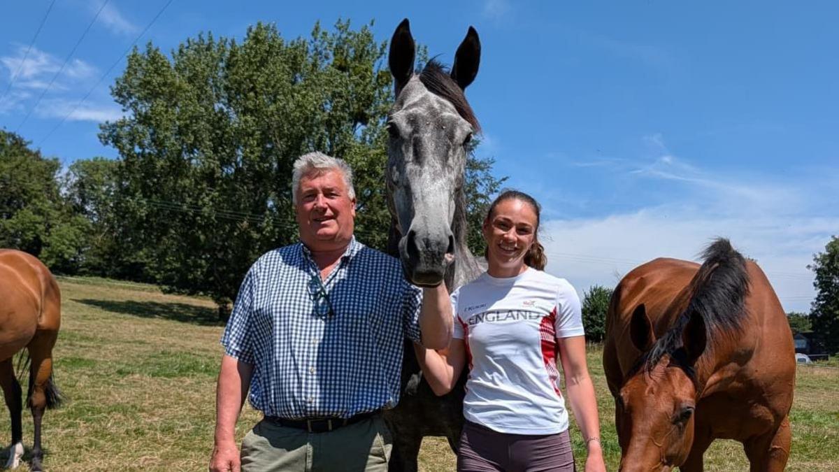 Paul Nicholls stands with his left arm holding the reigns of a grey horse in between Ellen Barber, wearing a white England Commonwealth Games T-shirt