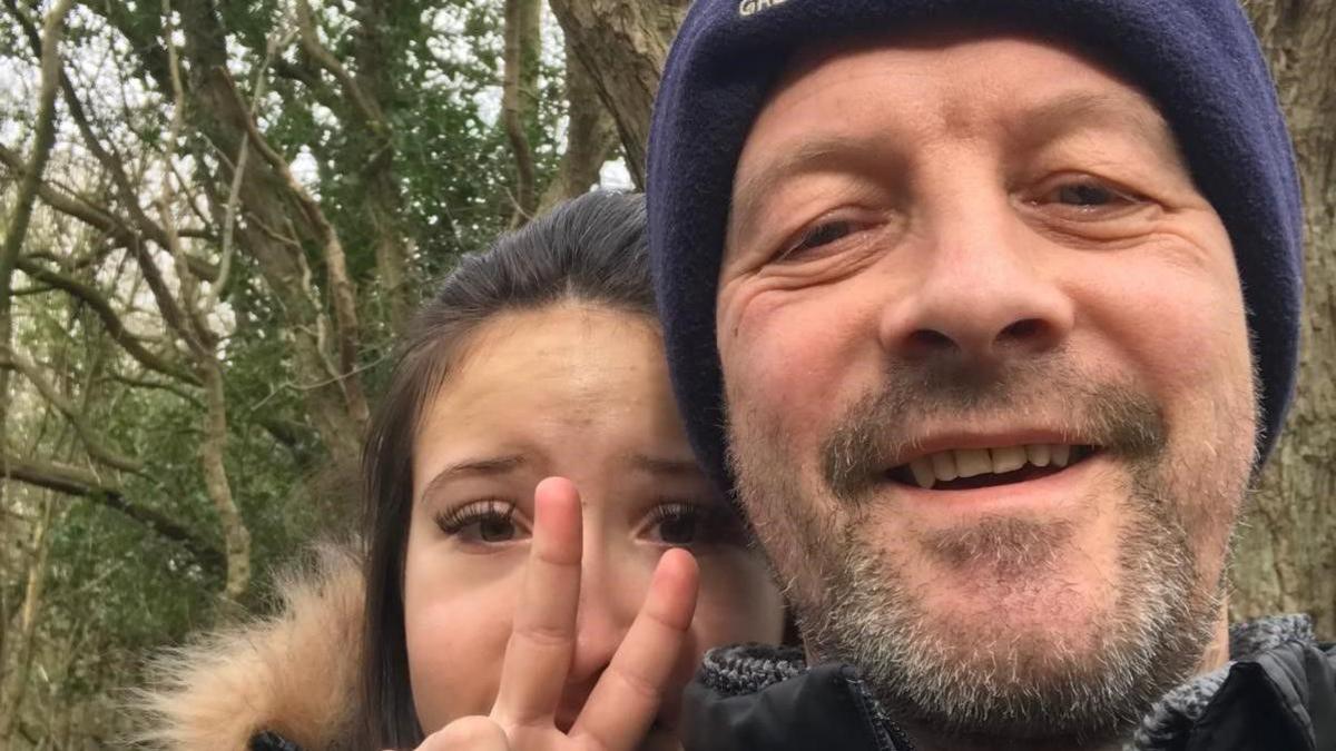 A father and daughter look into the camera. The dad is smiling and has stubble and a navy blue had on. The daughter is a teenager with dark hair, she is doing a 'peace' sign to the camera.