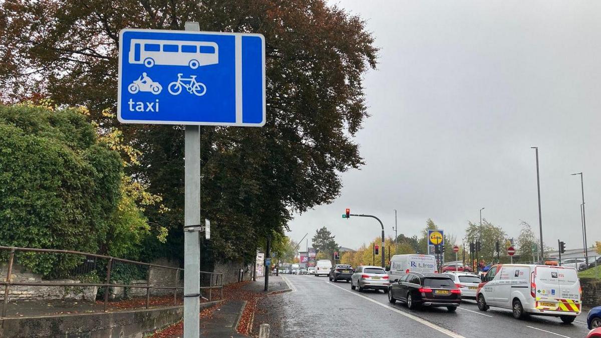 A blue road sign showing a taxi, motorbike, cycle and bus lane next to the road. There are lots of cars and work vans stopped at red lights, with a Lidl and wider retail estate shown in the background.