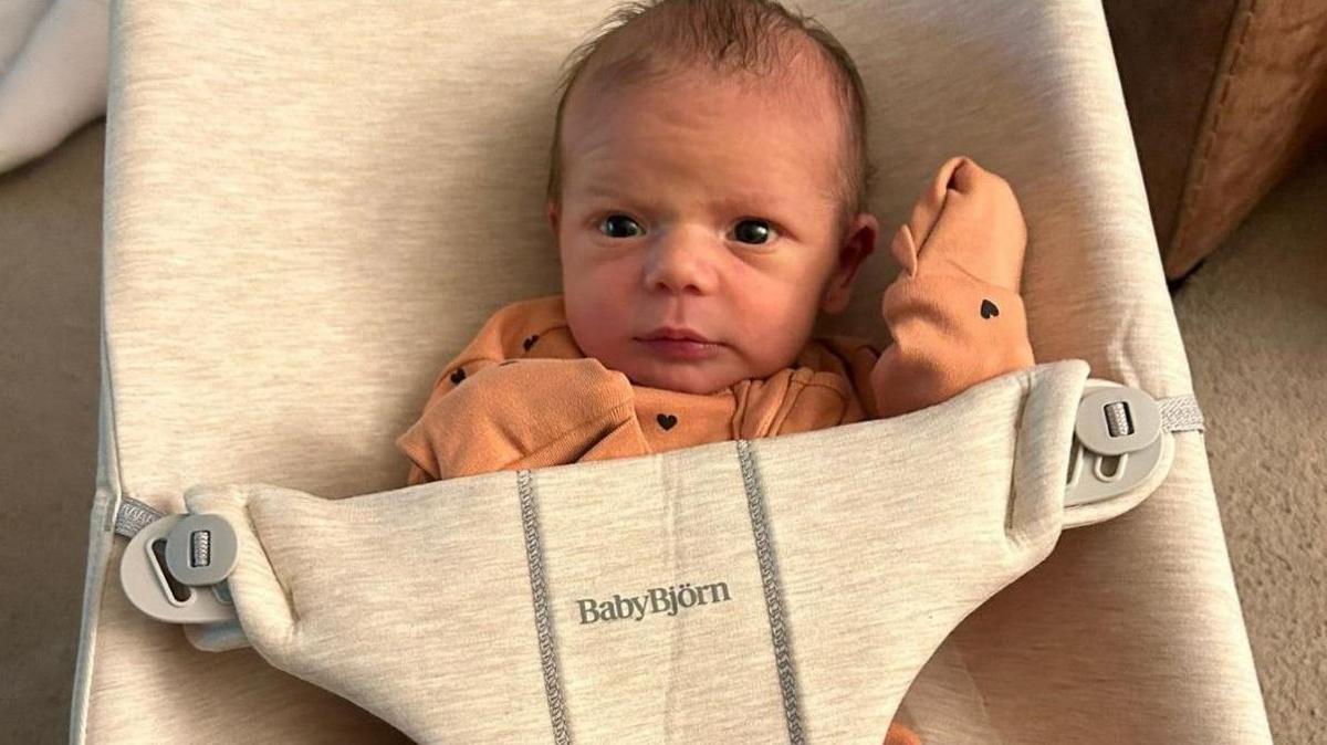 Baby in a little orange suit looks up towards camera as it sits in a fabric rocker chair