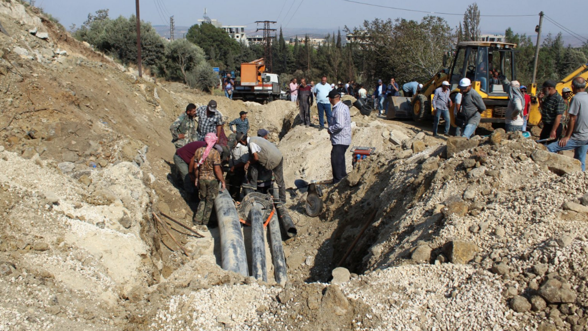 People inspect a damaged area in the aftermath of what Syrian state media reported was an Israeli strike in Masyaf. A yellow digger can be seen at the back. People in rubble search through sand.