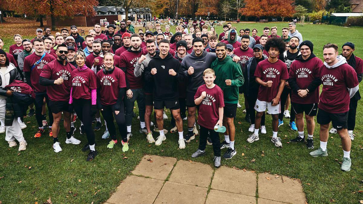 A large group of people dressed in running clothing stand in the middle of a park posing for a group photo. Most are wearing maroon t-shorts with "Matchroom Boxing" written on the front