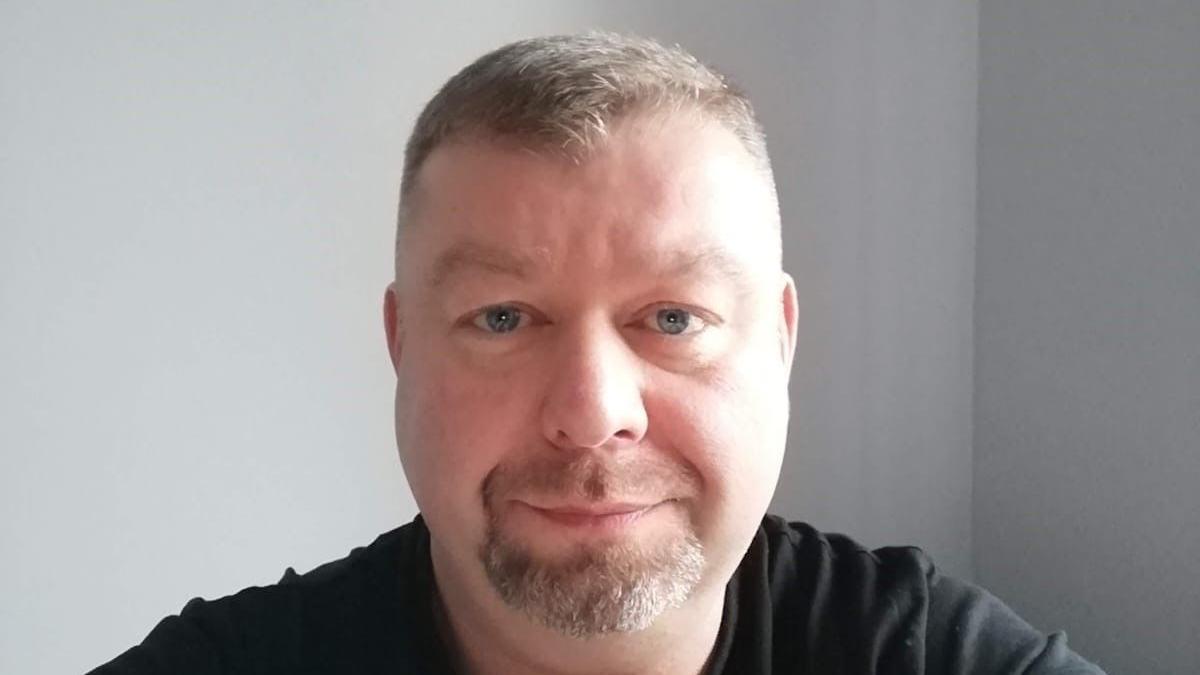A headshot of a man with short light hair and a goatee beard, He wears a black tshirt and stares into the camera.  