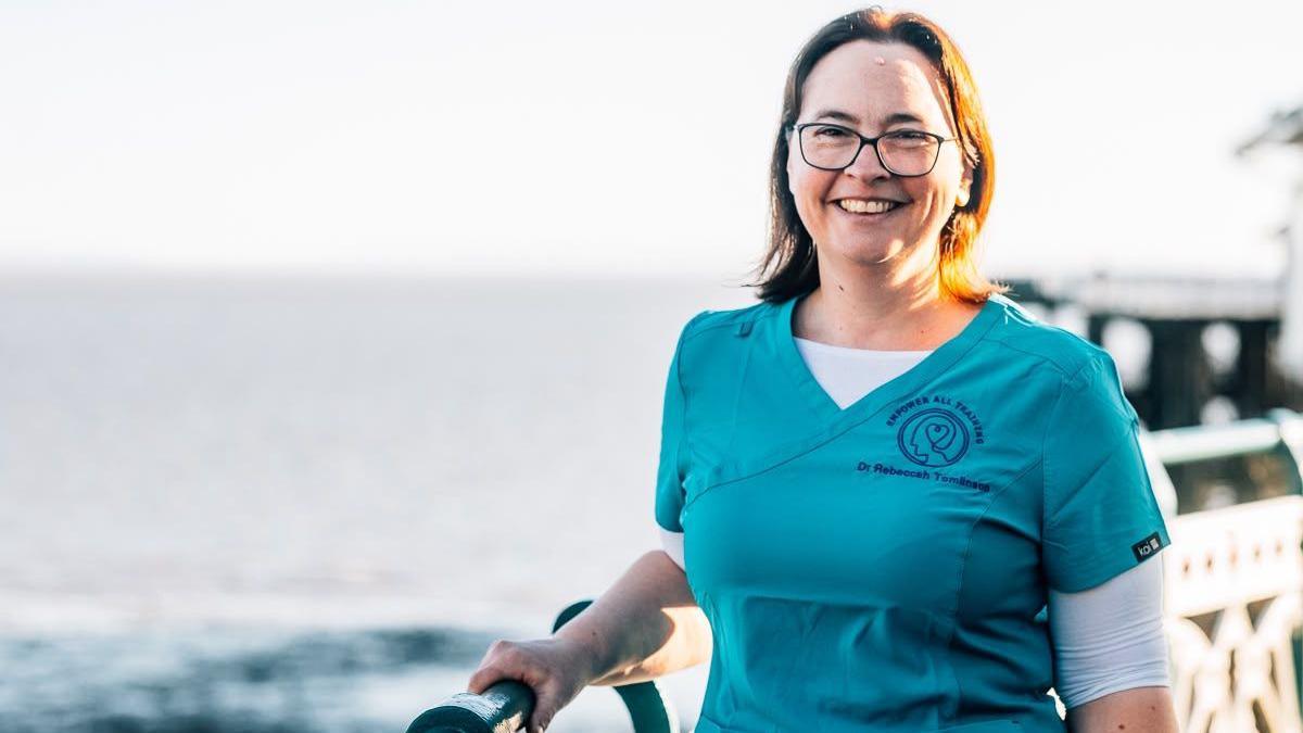 Dr Rebeccah Tomlinson mewn dillad meddygol glas yn sefyll ar bier gyda'r môr y tu ôl iddi