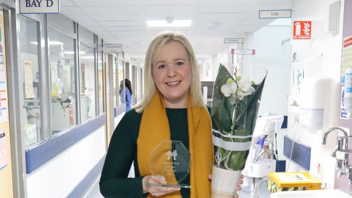 Young woman holds a glass sculpture and a pot of white flowers. A hospital ward is behind her.