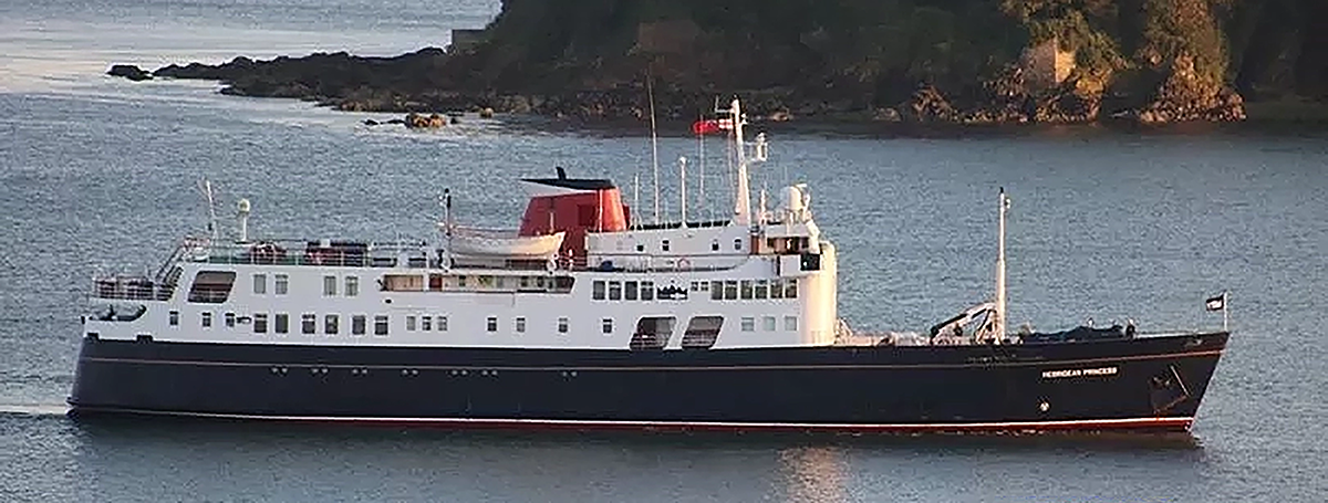 A small ship with black hull, white superstructure and red funnel at sea with rocky shore behind and warm sunset light.