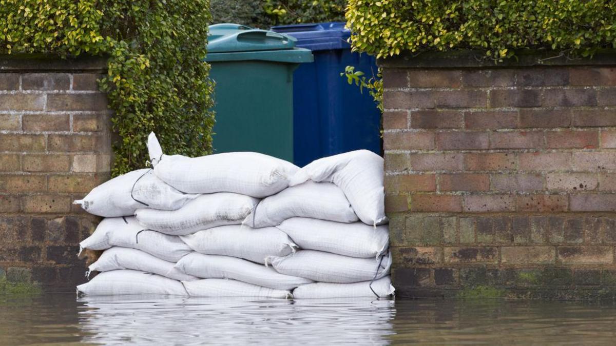 Sandbags are piled in the entrance way to a house. Brick walls are on either side, flood water is in front of the barrier.