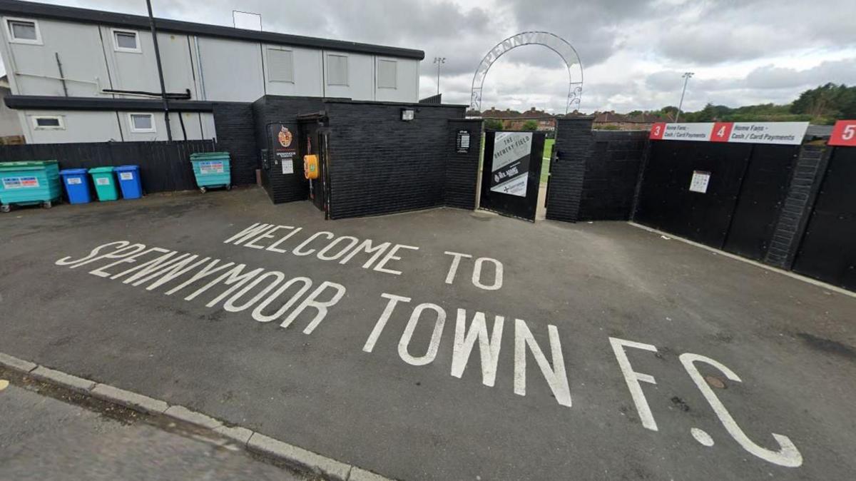 A sign reads "Welcome to Spennymoor Town FC" on the pavement outside the club's ground. A black brick wall surrounds the pitch and a white arch bearing the club's name stands above the entrance. A two-storey light grey building is on the left and closed turnstiles on the right.