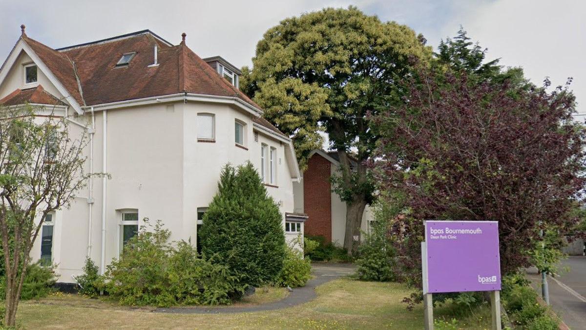 A Google Street View image of a large house on a corner plot, set in a lawned garden lined with trees and shrubs. Near the boundary is a large purple sign which says BPAS Bournemouth