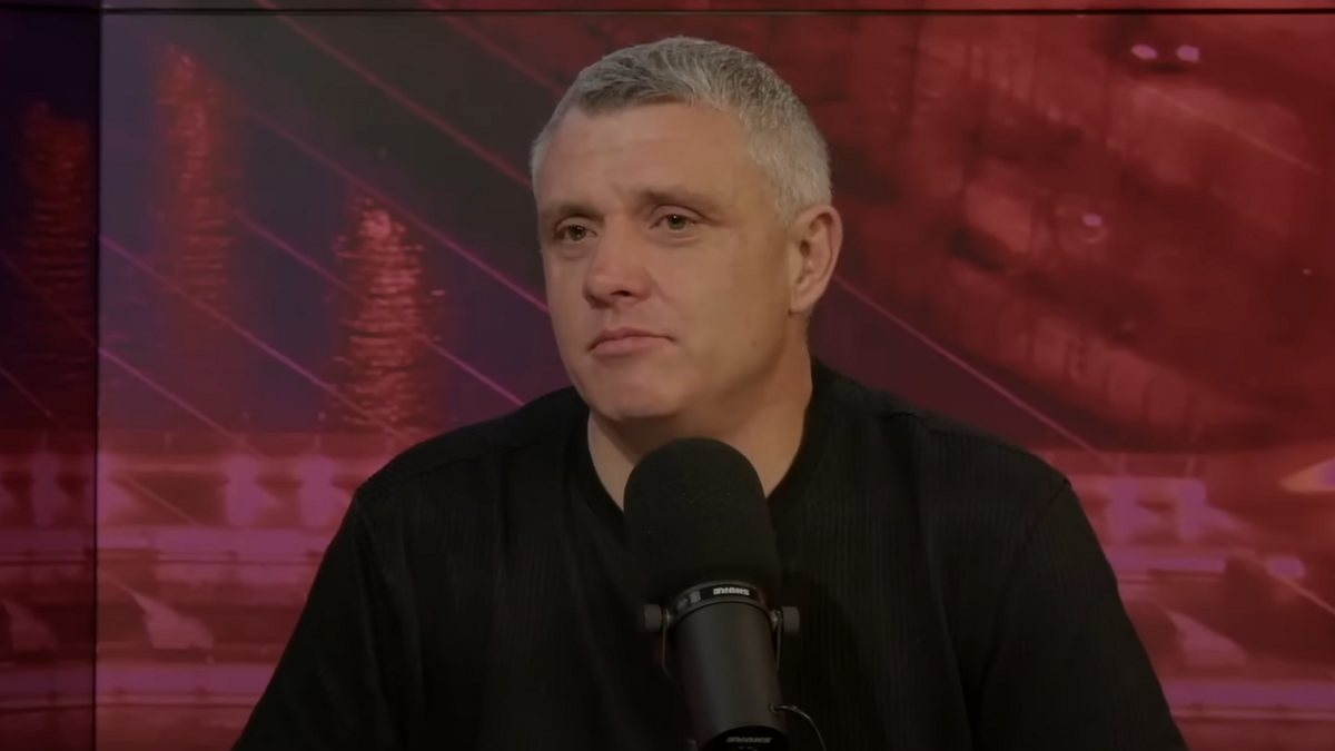 A screenshot of Niall Donald sitting in front of a black, table microphone while recording the Crime World podcast. Mr Donald is an older man with short, greying hair and is wearing a black t-shirt. He is sitting in front of red screen, displaying the Samuel Beckett bridge.