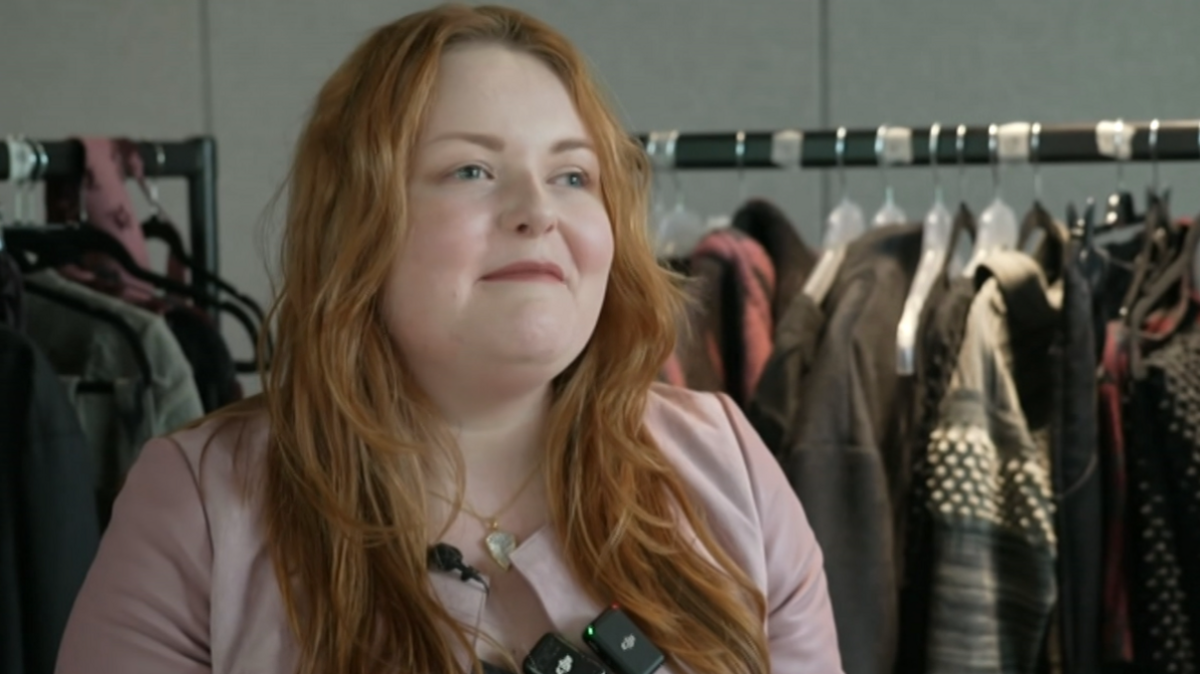 Lucy Edwards, wearing a pink top and a necklace, in front of a rack of clothes. She has long strawberry-blonde hair, blue eyes and has a slight smile