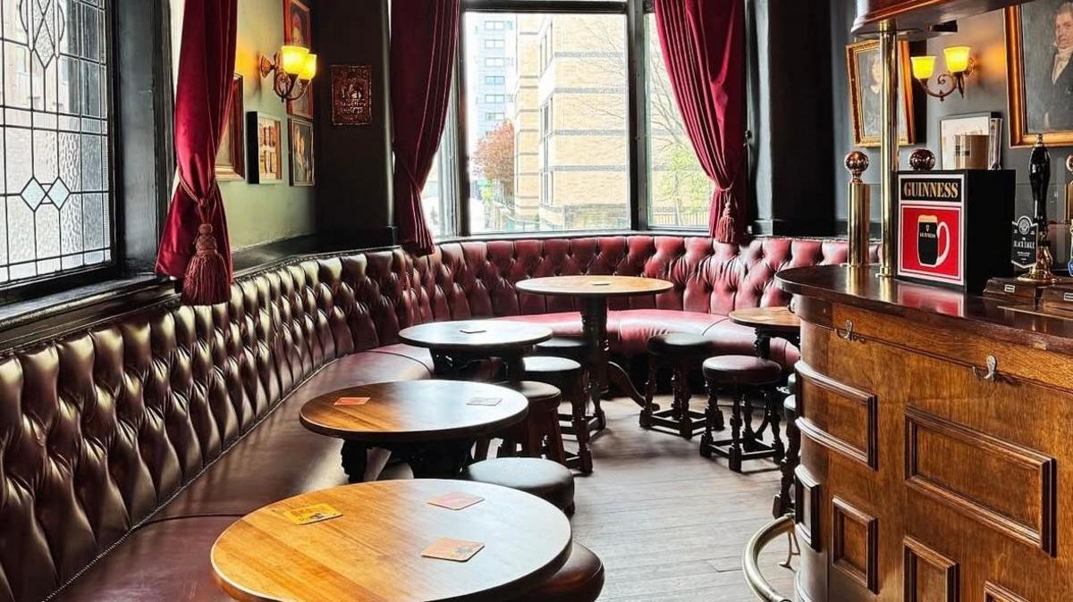 Interior of a traditional British pub with a long, tufted leather banquette with paired with round wooden tables and stools. Large windows with stained glass panels and heavy red curtains let in daylight. To the right, there is a wooden bar with a brass rail and a prominent Guinness sign.