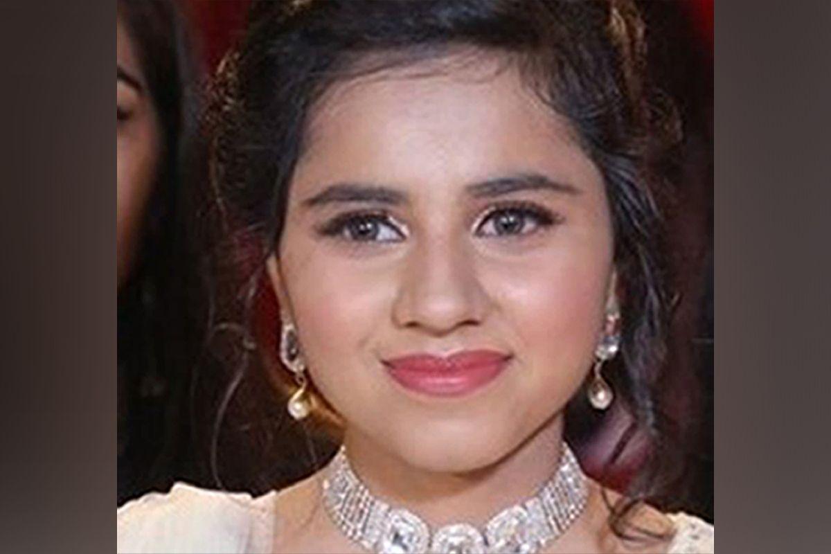 A close up photo of Harshita Brella wearing a white dress, a diamond looking necklace and gold earrings and red lipstick. She is smiling without teeth