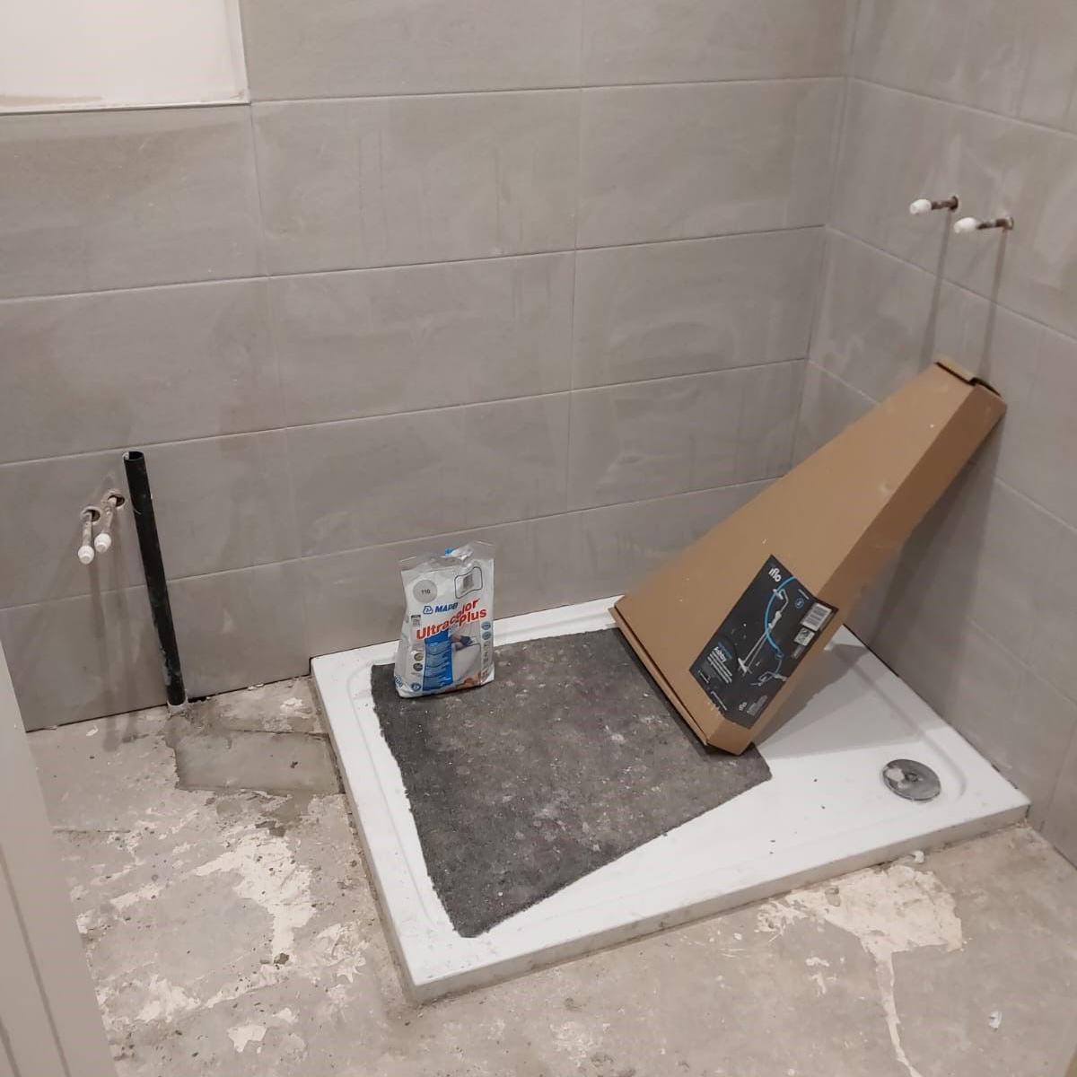 Unfinished bathroom with concrete floor, shower tray, but no shower cubicle, and tap points coming out of the wall, but no taps. The walls are tiled a grey colour that looks unfinished. The shower tray has a cardboard box on it, and a packet that looks like grout or other building materials, along with a section of black matting or underlay.
