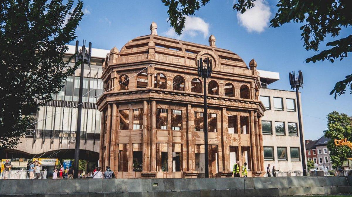 An artist's impression of a large cardboard building, almost matching the height of the three-storey buildings behind it. The structure is on a city square, with trees visible on either side.