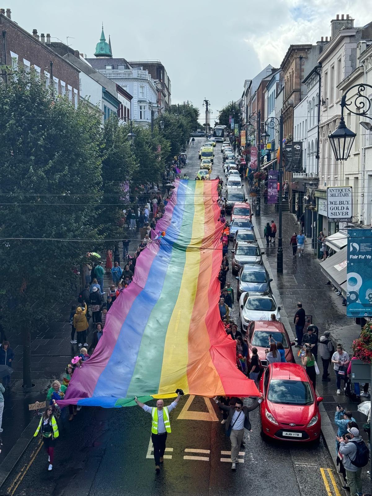 Large rainbow Pride flag being carried down the length of an entire road in Derry