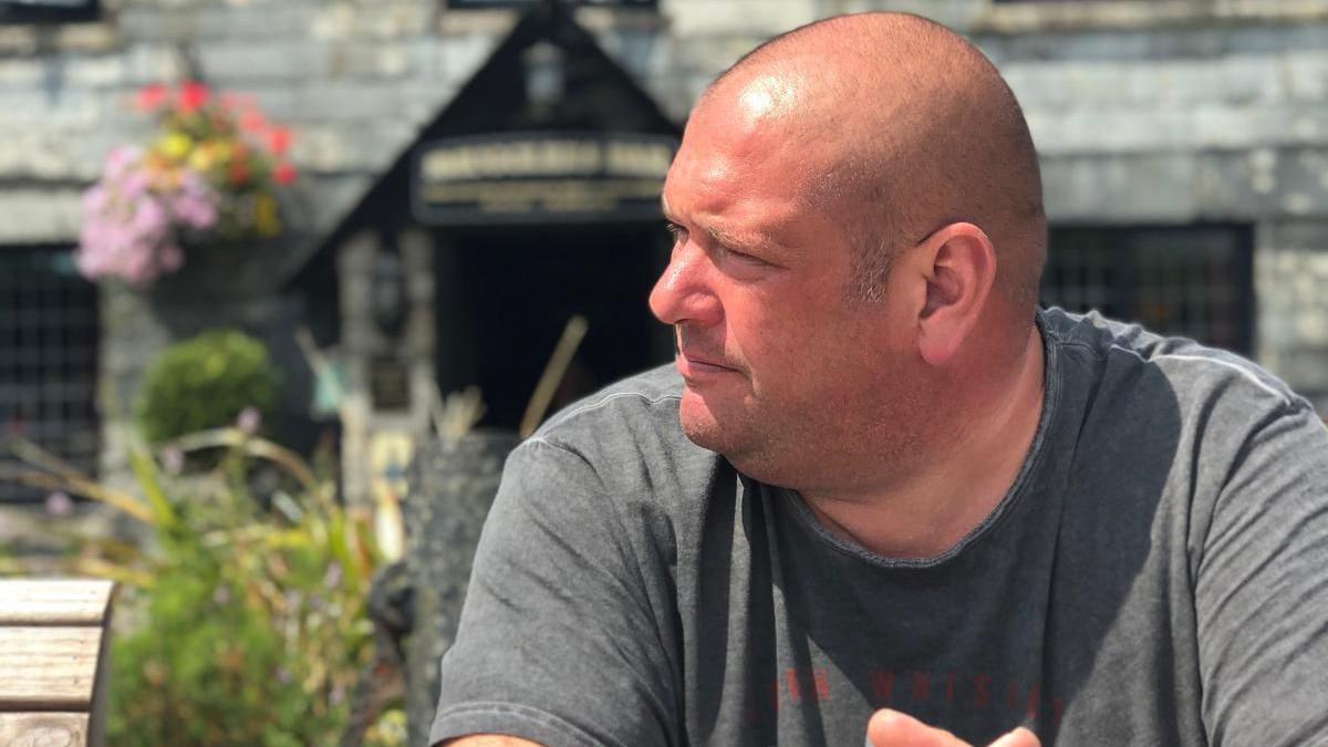 Image of Mark in a beer garden. His left side facing the camera. It is sunny in the picture.
