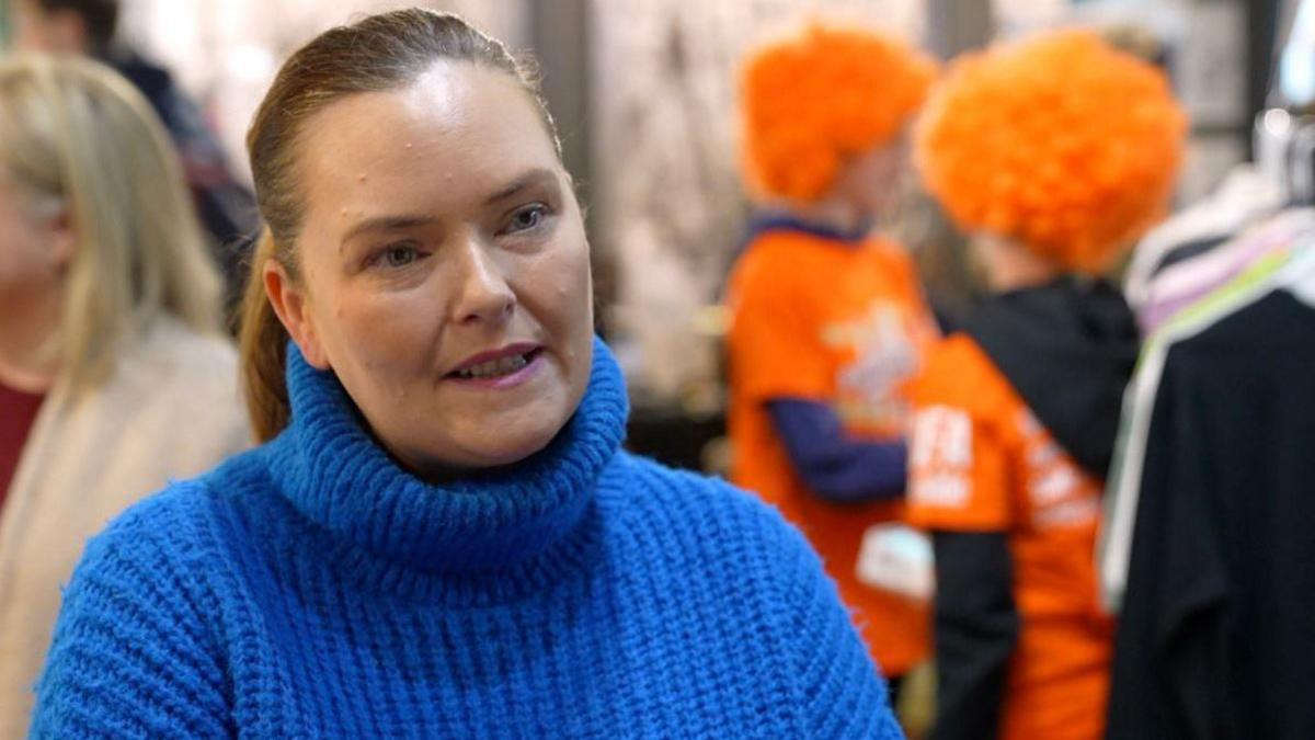 A woman in a bright blue woollen polo neck jumper. She has fair hair tied back in a ponytail. In the background, figures in orange t-shirts and orange curly wigs can be seen.