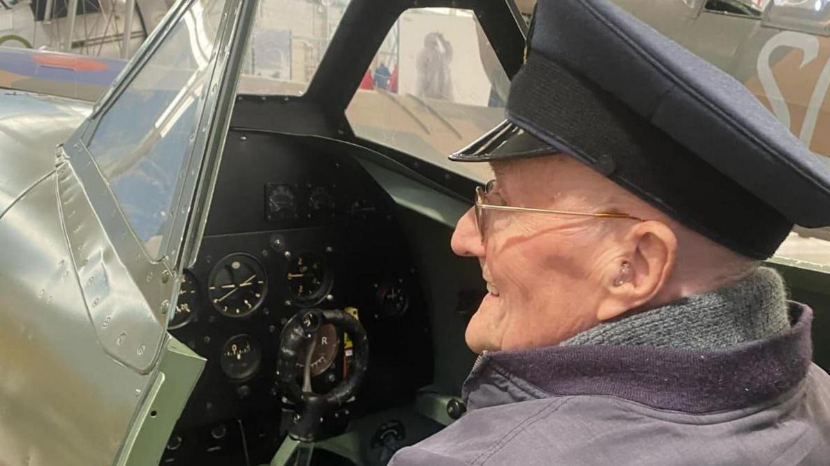 An over-the-shoulder view of John sitting in the cockpit. He is wearing a peaked cap and glasses and is smiling, with the aircraft's instrument display in front of him.