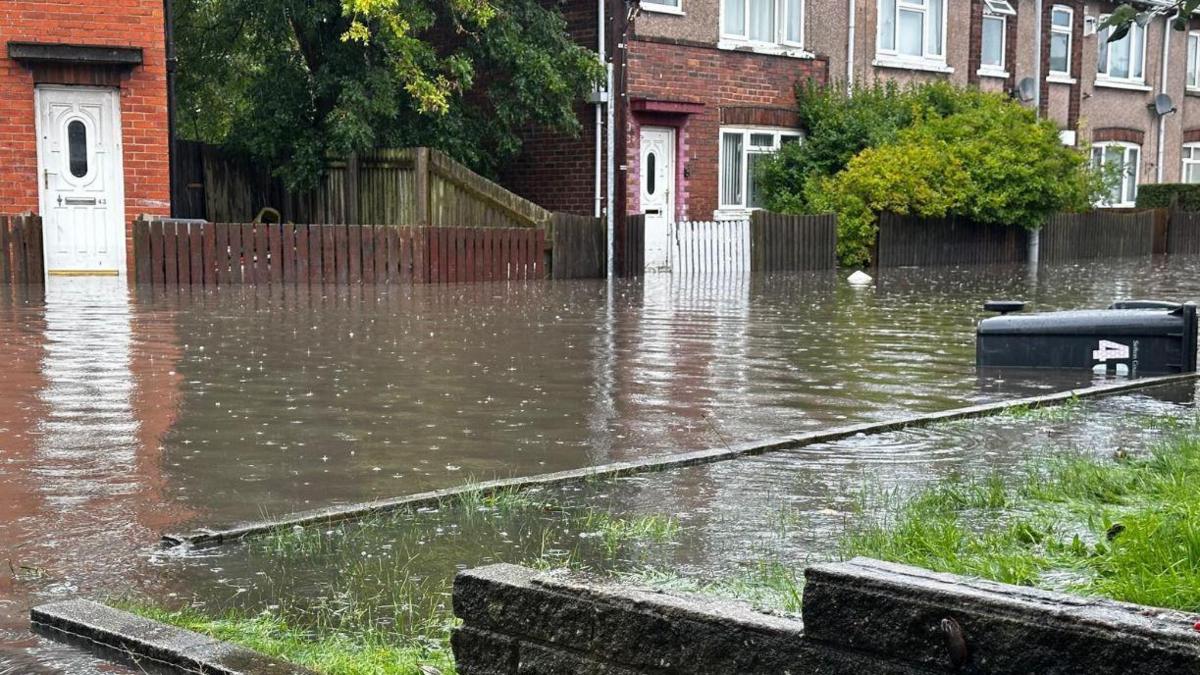 A flooded terraced street with water up to the door frames and a wheelie bin lying on its side partly submerged in water