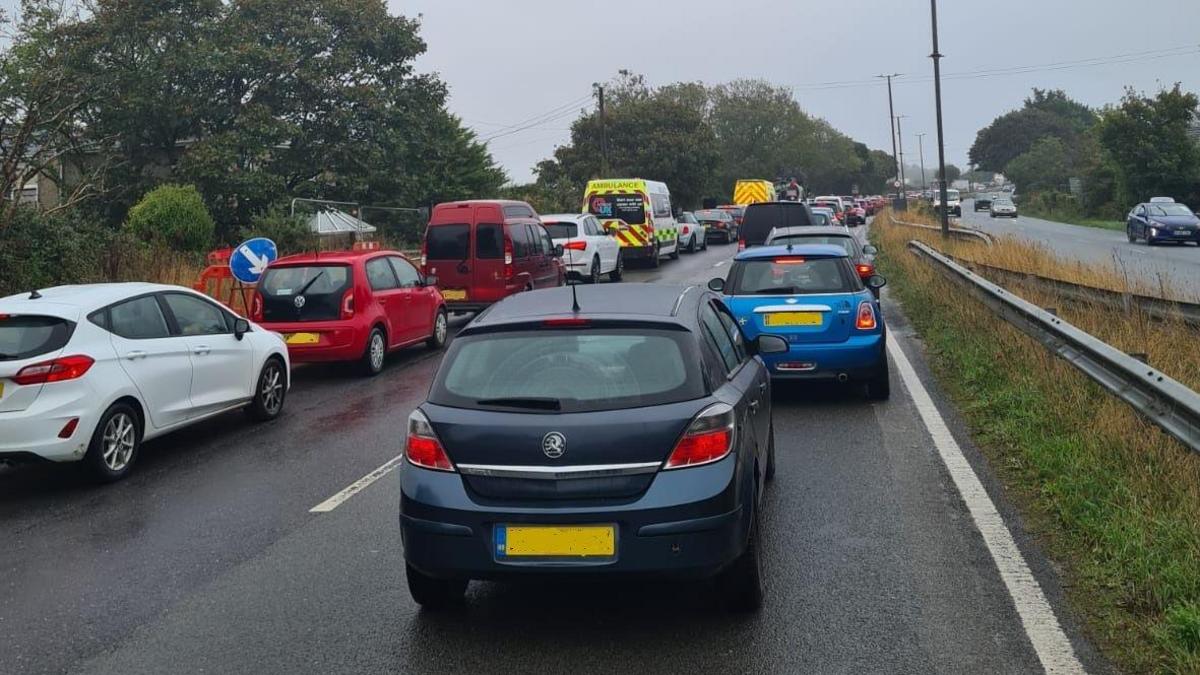 Cars are stationary and queued up on the A390 dual carriageway. An ambulance is among them. The lanes in the opposite direction are relatively empty. It is a grey day.