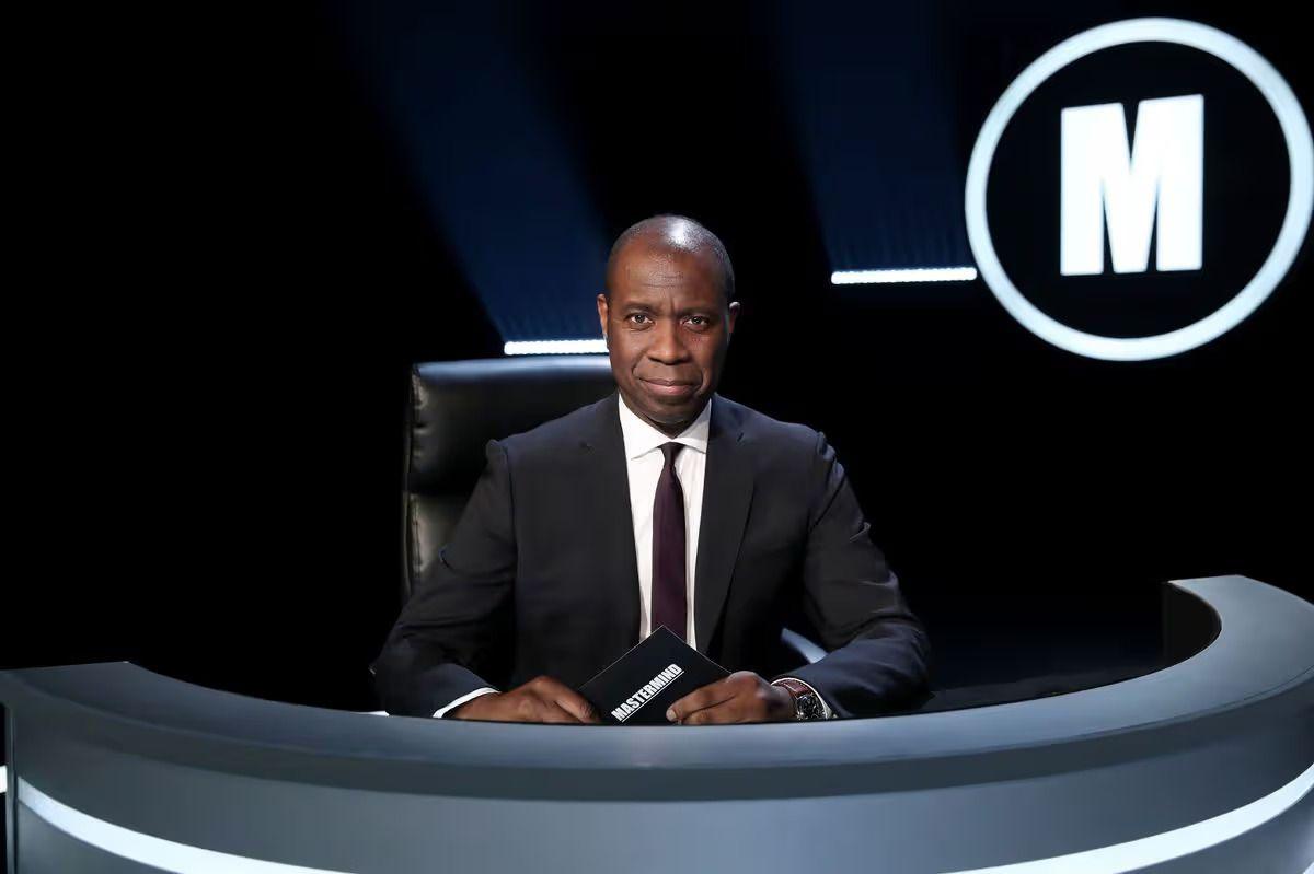 clive myrie, a man in a black suit with a dark red tie, sits behind a curved black desk. in his hand is a black card with the word mastermind written on it