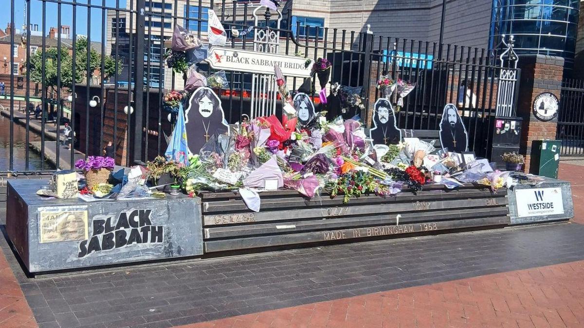 A bench with black and white images of the members of Black Sabbath has flowers resting across the bench