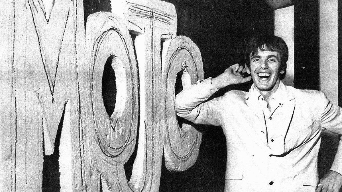 A black and white photograph of a man who is smiling as he leans with his elbow against a sign that reads "Mojo".