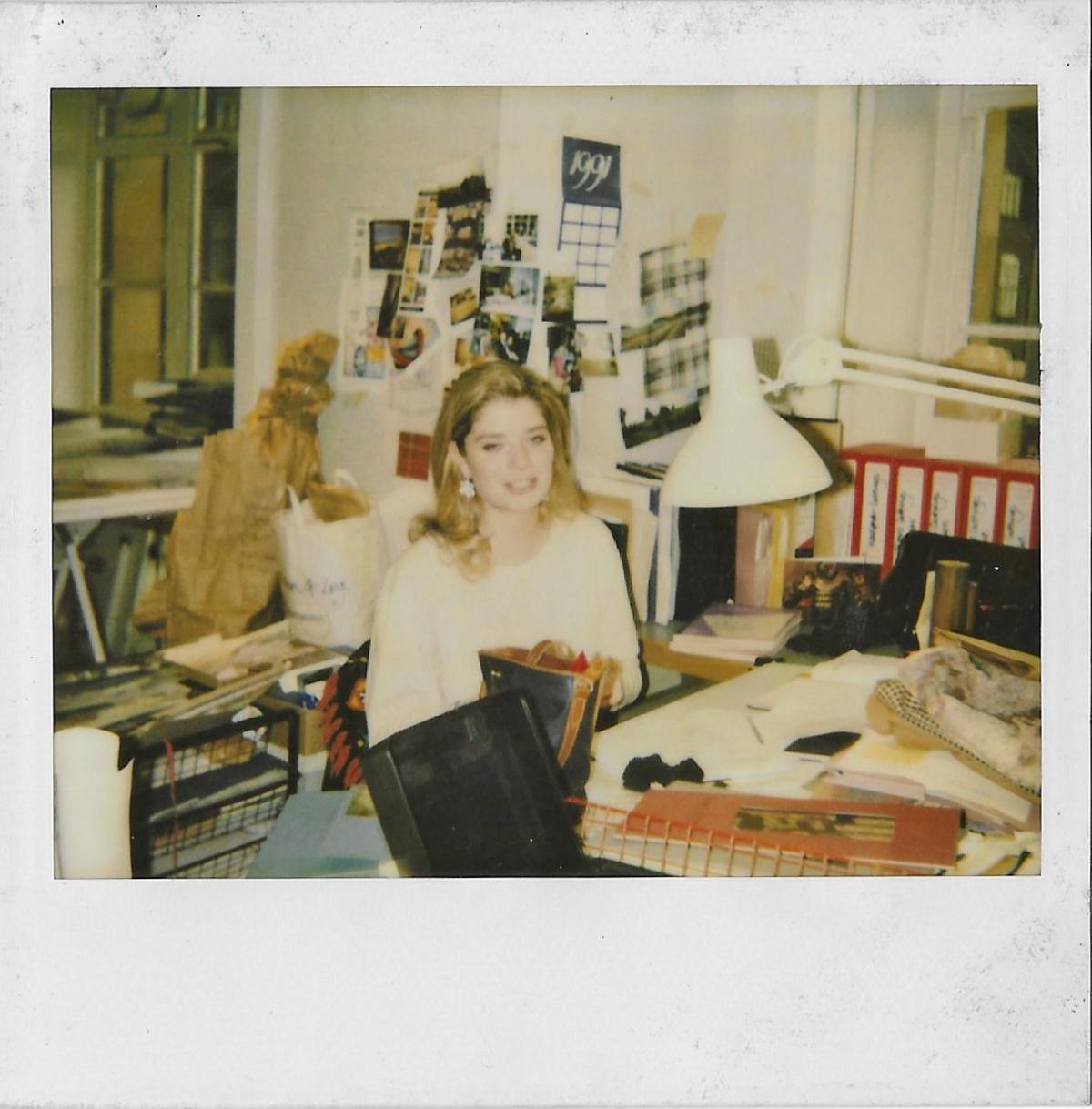 A polaroid picture taken in 1991 of Frances inside the Harrods office. She is smiling and sitting behind a desk busy with papers and books. Behind her are red files and a notice board with a calendar, pictures and more papers attached.  